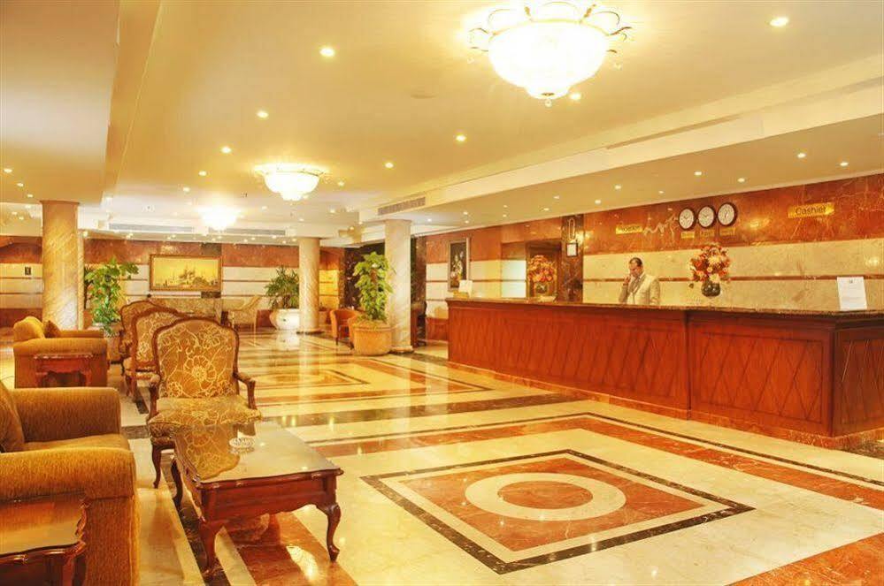 Sunrise Alex Avenue Hotel Alexandria Exterior photo The photo shows a hotel lobby with an elegant design. There is a reception desk made of polished wood, featuring decorative elements and a few clocks. The floor displays a striking marble pattern with circular designs. On either side of the lobby, th